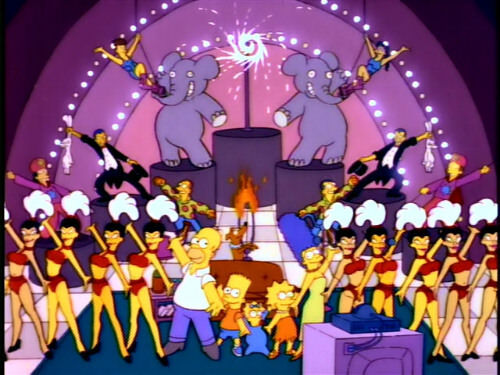 The iconic couch from the Simpsons show appears on stage in a flashy vaudevillian act surrounded by show girls with headpieces.