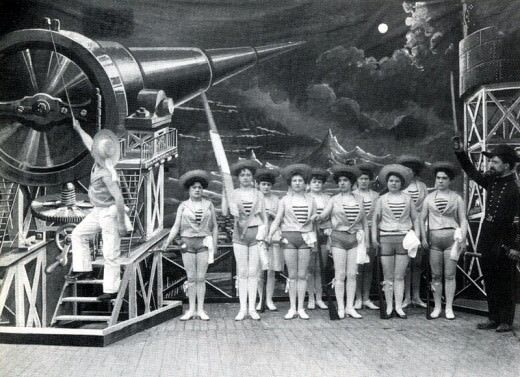 Still from the black and white movie A Trip to the Moon. We see a cast of females in shorts on stage, and a man who is dressed like a sailor descending a ladder. At the top of the ladder is a futuristic machine pointed at the moon.