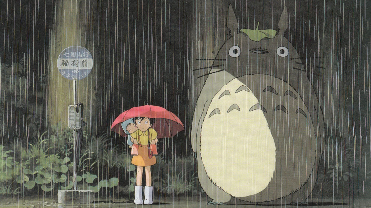 Totoro, a large fictional animal stands in the rain at a bus stop next to a person holding a red umbrella.