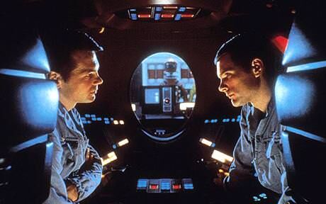 Within a dark interior of the spaceship, two astronauts face each other, looking concerned.