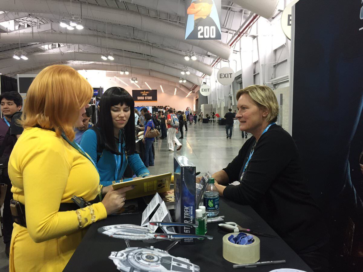 Denise Crosby sits at a booth at the New York Star Trek Convention across from two fans. She is signing autographs.