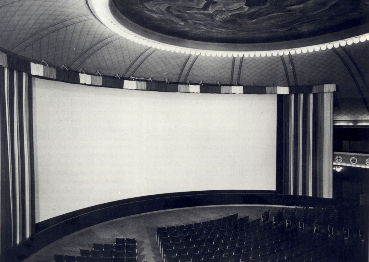 This black and white photograph depicts the interior of a Cinerama movie theater with the curved screen. The lights are on, and there is no one in the audience.