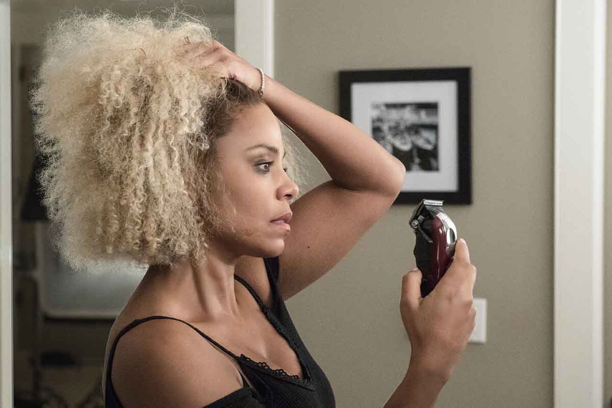 A woman with long hair looks into the mirror with a shaver in hand.