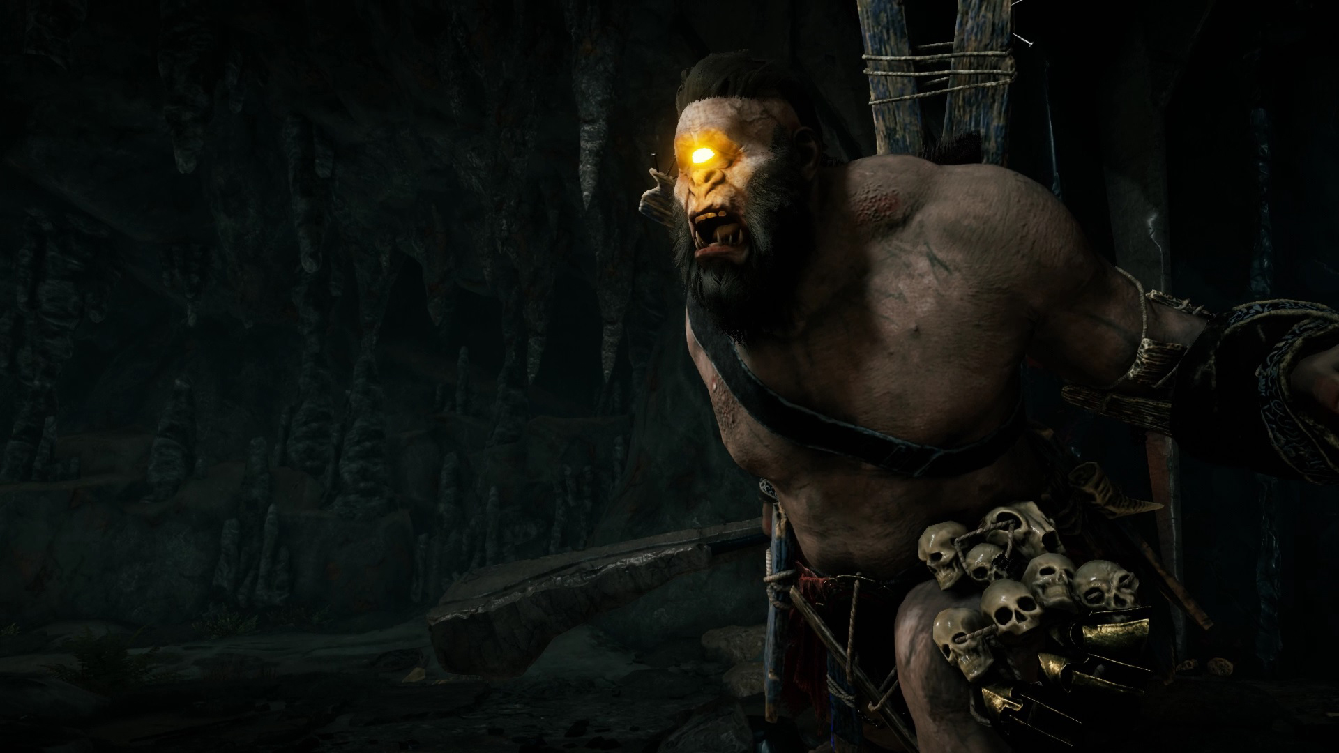O Ciclope Brontes, à direita da imagem, no interior de uma caverna escura. Seu único olho brilha feito um farol amarelo. O rosto e a postura são ameaçadores. Ele carrega no cinto vários crânios humanos.