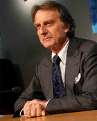 Upper body of a man in his sixties with neatly combed, brown but greying hair, wearing a smart grey suit and navy tie with small white spots. He is sitting at a table, upon which his hands rest.
