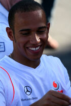 Head and shoulders of a man in his twenties, who has close-cropped black hair and is showing his teeth as he smiles while signing something unseen. He is wearing a white, V-neck T-shirt displaying the Mercedes-Benz and Vodafone logos.