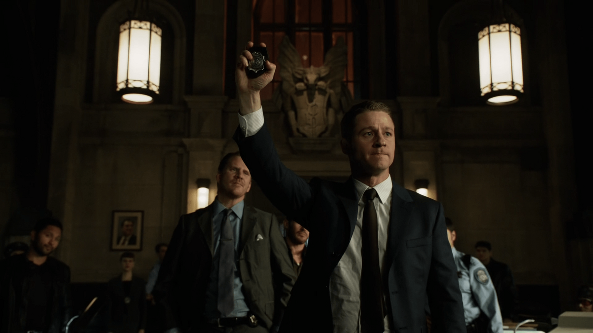 Detective Jim Gordon is seen center-screen, flanked by his colleagues inside a dark, gothic interior. He is walking forward authorotatively, with his hand up in the air, displaying his badge.