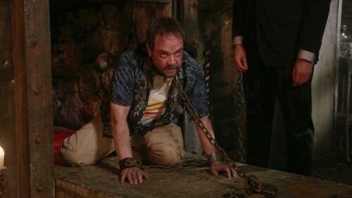 Crowley is seen on his knees with a chain around his neck that is attached securely to the floor.