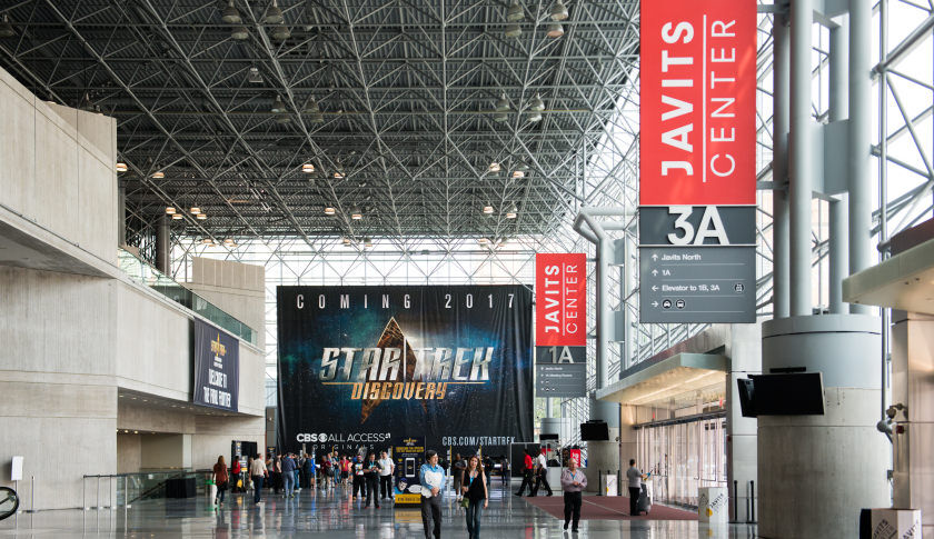A view inside the front hallway of the Jacob Javits Center convention building for the Star Trek: Mission New York convention. Convention-goers mill about. A bill-board sized banner depicting the Star Trek Coming in 2017 logo hangs on the far wall.