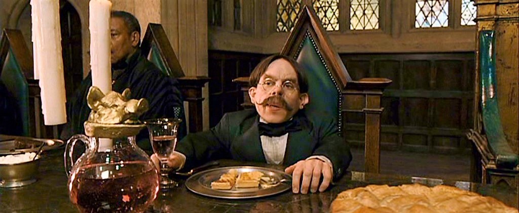Profesor Kratiknot sedící na zeleně polstrované židli s rukama na stole, v jedné drží pohár s nápojem.