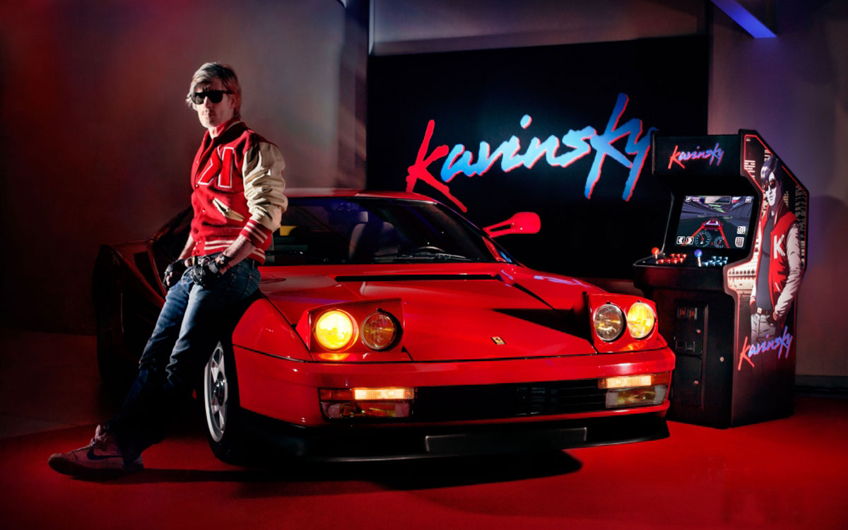 An image of French DJ, Kavinsky, leaning on his iconic, red, Ferrari Testarossa, with a Kavinsky branded arcade cabinent behind him