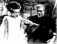 The Bride of Frankenstein has black hair with a white streak running through it, is dressed in a white gown, and has a blank expression. She is standing on the left with her left hand elevated. On the right is Frankenstein's monster, standing on the right and smiling. His right hand is below hers. The background includes walls made of stone.