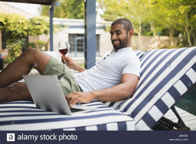 Smiling-man-holding-wineglass-while-using-laptop-on-lounge-chair-at-J84X33